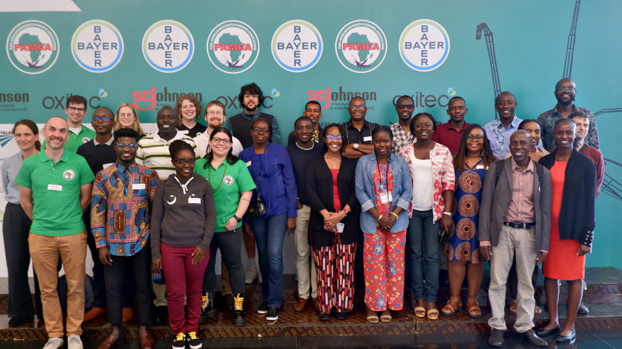 A group of people standing together and smiling. Some people are wearing matching green t-shirts and all are wearing lanyards. They appear to be indoors, with a large blue banner behind them. The banner has several logos on it, including PAMCA and Bayer.