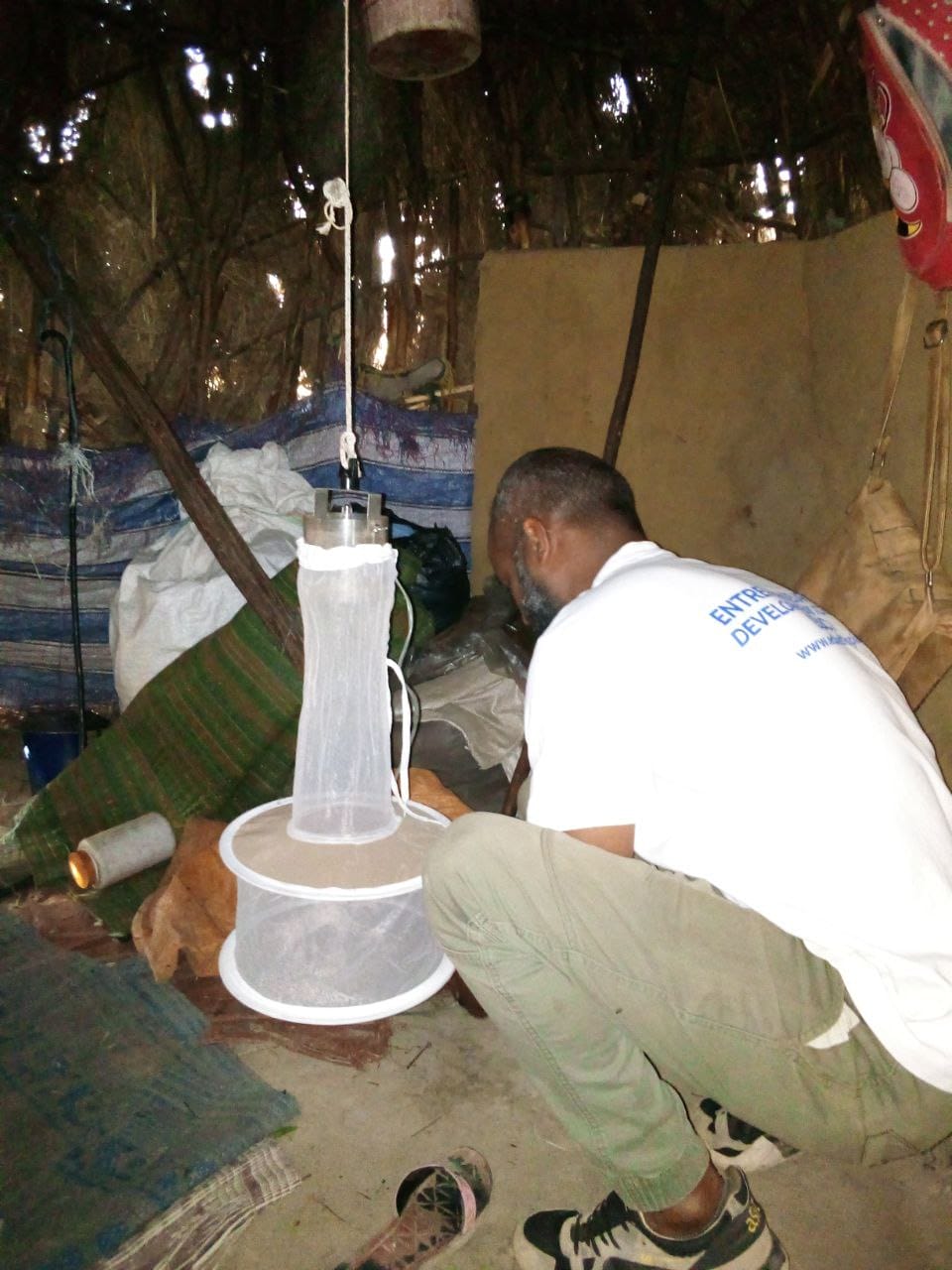 A person wearing a white t-shirt squatting down inside a cluttered room, and focused on a mosquito light trap in front of them.